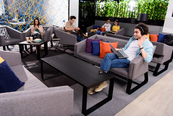young businessman in headphones sitting on couch with laptop and waving by hand at modern coworking office  - Foto, afbeelding