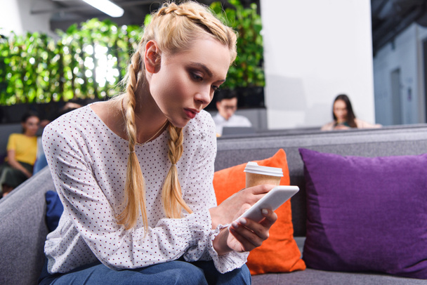 junge Geschäftsfrau mit Pappbecher Kaffee und Smartphone auf Sofa im modernen Coworking-Büro  - Foto, Bild