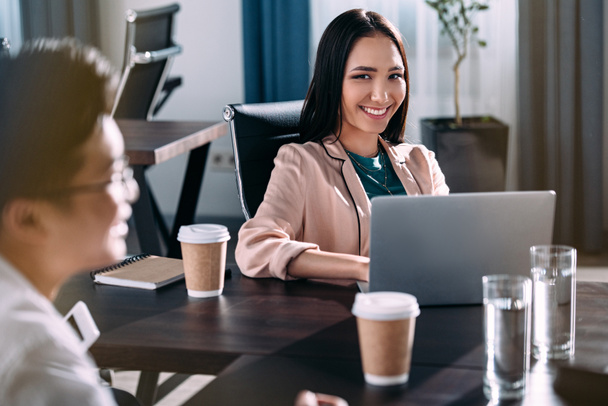 молода азіатська бізнес-леді сидить за столом з ноутбуком, кавою і водяними окулярами, а її партнер обговорює в сучасному офісі
  - Фото, зображення