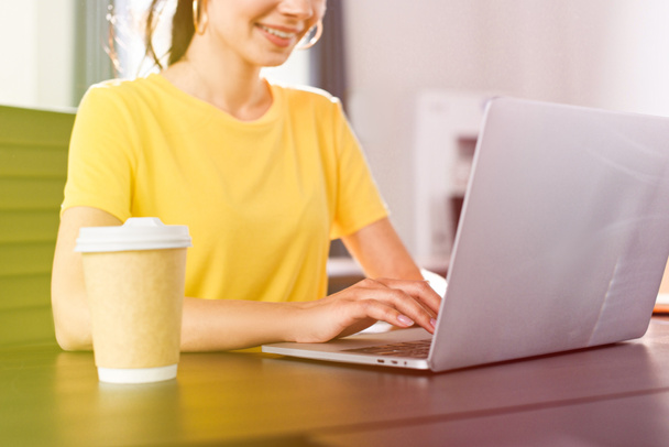 bijgesneden afbeelding van Glimlachende zakenvrouw zittend aan tafel met laptop en koffie kopje in moderne kantoren  - Foto, afbeelding