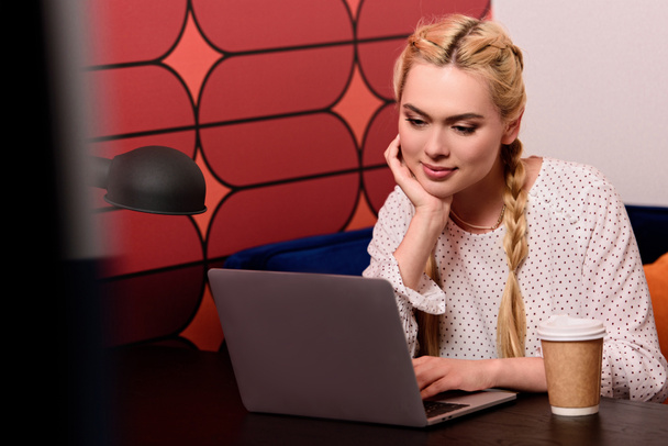 young businesswoman with paper cup of coffee sitting at table with laptop in modern office  - Foto, Bild