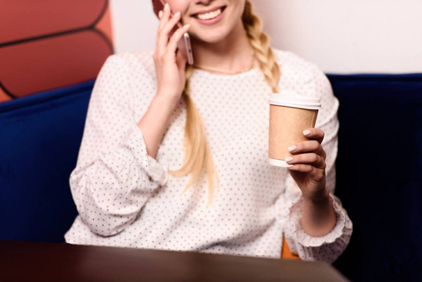 cropped shot of smiling businesswoman talking on smartphone and drinking coffee in office  - Zdjęcie, obraz