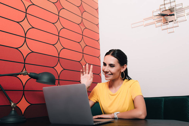 laptop ile masada oturan ve ofiste el sallayarak genç iş kadını  - Fotoğraf, Görsel