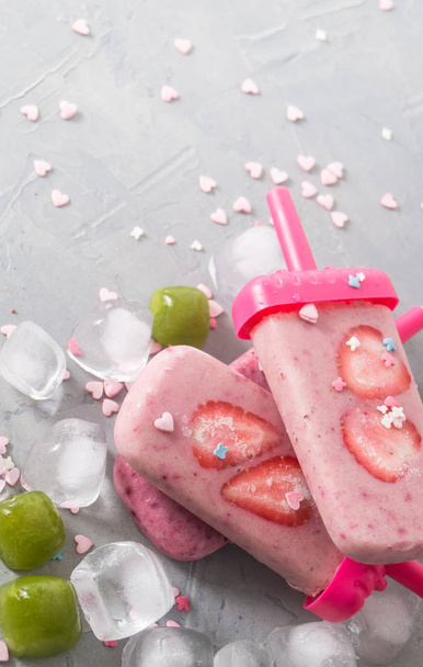 Summer heat: refreshing pink fruit ice cream with strawberry pieces surrounded by transparent and green ice cubes on a gray table. - Photo, Image