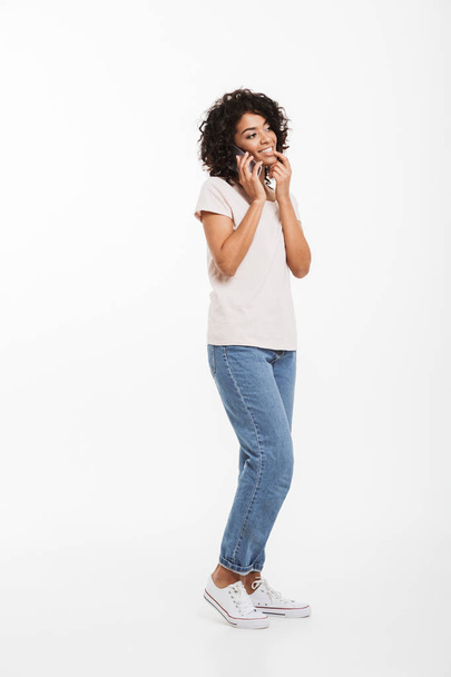 Full length picture of content american woman with afro hairstyle wearing t-shirt and jeans speaking on cell phone with pleasure isolated over white background - Foto, immagini