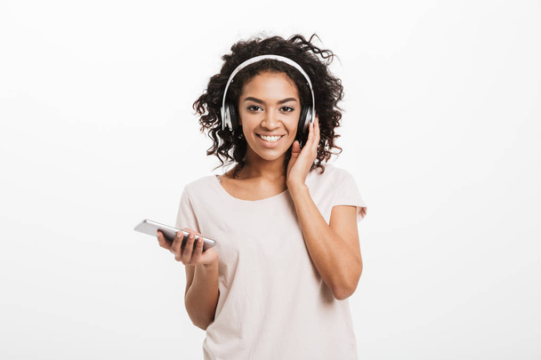 Fascinating american woman with curly hairstyle and big smile listening to music via wireless earphones holding silver smartphone in hand isolated over white background - Foto, Imagen