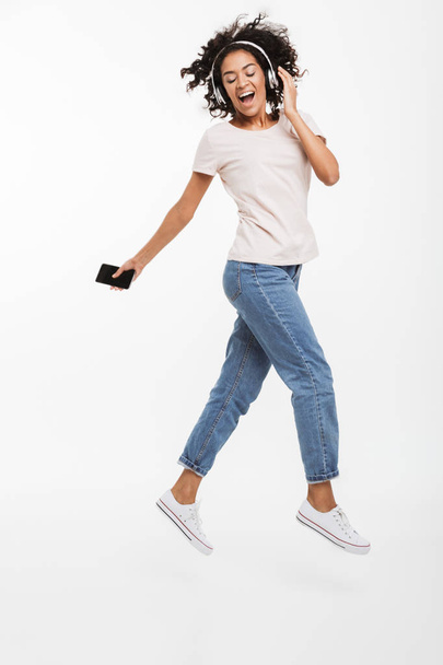 Immagine a figura intera di donna afroamericana soddisfatta che prova piacere mentre ascolta musica utilizzando cuffie wireless e smartphone isolati su sfondo bianco
 - Foto, immagini