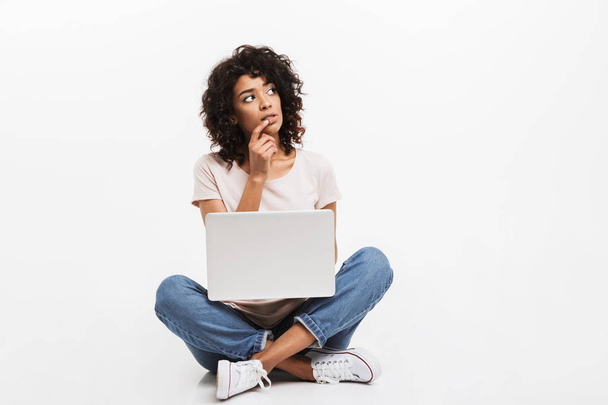 Portrait of pensive young afro american woman using laptop while sitting on a floor with legs crossed isolated over white background - Fotoğraf, Görsel