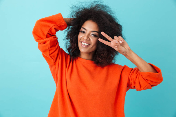 Photo of american woman in red shirt looking on camera and showing peace sign isolated over blue background - Fotografie, Obrázek
