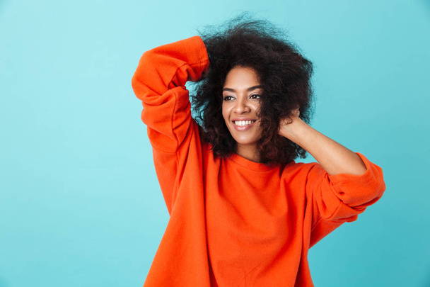 Colorful portrait of american woman in red shirt looking aside with smile and touching her messy hair isolated over blue background - Zdjęcie, obraz