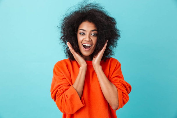 Colorful portrait of excited woman in red shirt looking on camera with smile and raising hands to face isolated over blue background - Foto, imagen