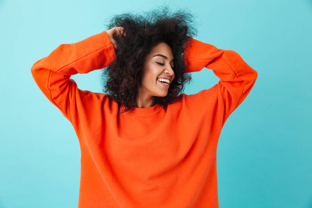 Colorful portrait of smiling woman in red shirt looking aside and touching her dark curly hair isolated over blue background - Фото, изображение