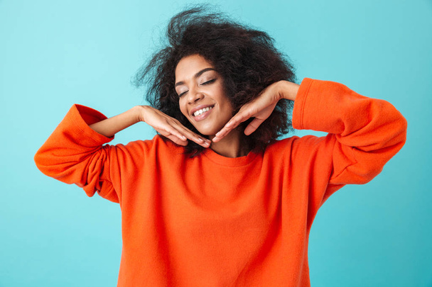 Young american woman in orange shirt posing on camera with lovely smile and keeping hands at face isolated over blue background - Φωτογραφία, εικόνα
