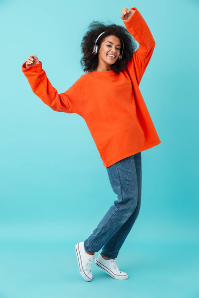 Full length photo of american smiling woman in orange shirt and jeans listening to music via wireless earphones isolated over blue background - Φωτογραφία, εικόνα