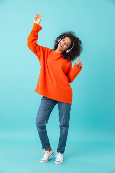 Full length multicolor photo of pleased woman in casual jeans dancing while listening to music using wireless headphones isolated over blue background - Foto, immagini