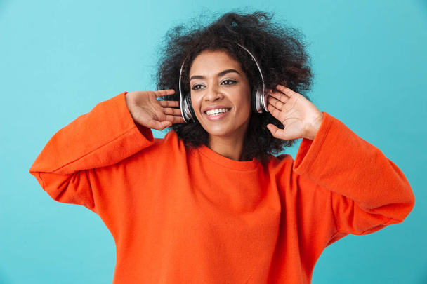 Pleased american woman 20s with shaggy hairdo enjoying music via wireless earphones while listening to favorite tune isolated over blue background - Foto, Bild