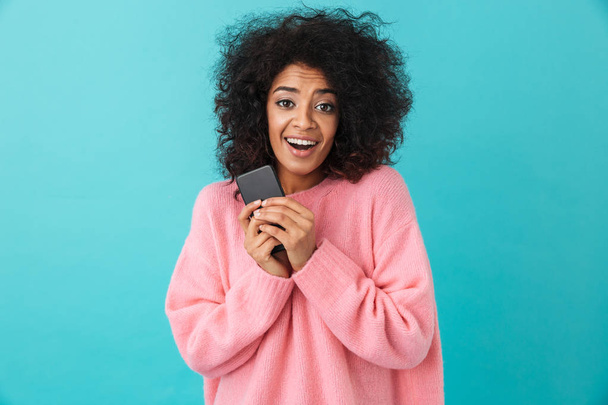 Image of american curly woman in pink shirt rejoicing and holding black mobile phone in hands isolated over blue background - Foto, Imagen