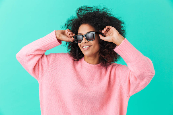 Fashion portrait of stylish woman 20s with afro hairstyle smiling and wearing black modern sunglasses isolated over blue background - Zdjęcie, obraz