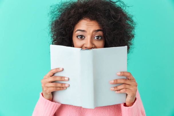 Positive image of american woman 20s with afro hairstyle looking on camera and covering face with interesting book isolated over blue background - 写真・画像
