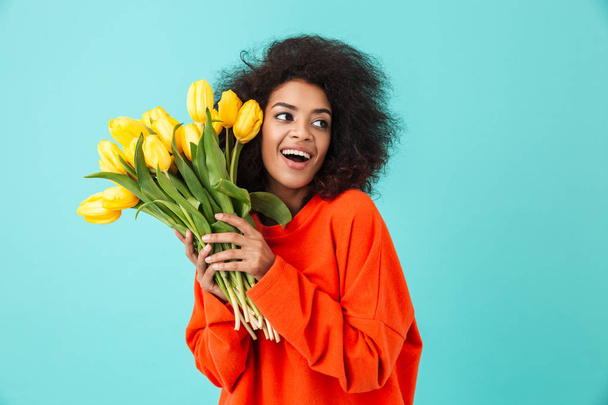 Curly american woman in red clothing looking aside and holding bunch of beautiful yellow tulips isolated over blue background - Фото, изображение