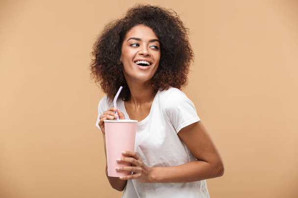 Portrait of a cheerful young african woman holding a plastic cup isolated over beige background - Foto, immagini