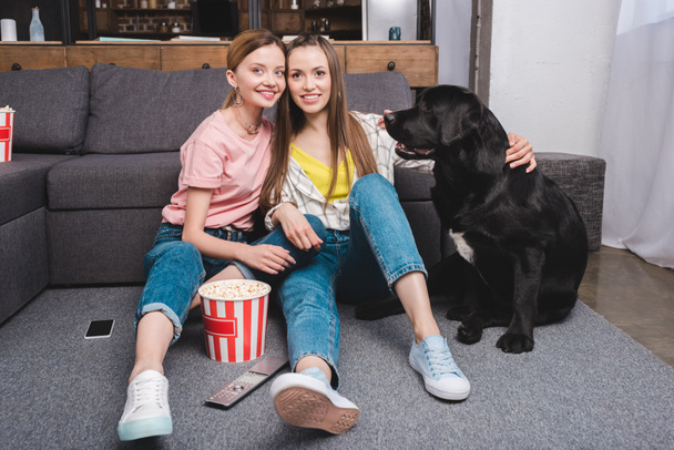 two smiling female friends with black labrador sitting on floor with remote controller, popcorn bucket and smartphone  - Foto, Imagem