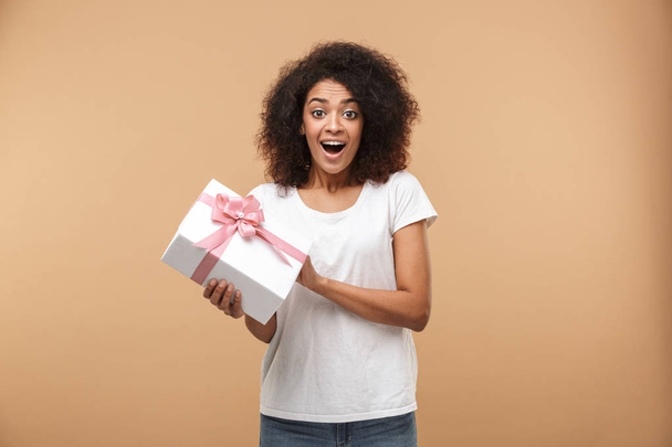 Portrait of an excited young african woman holding present box isolated over beige background - Valokuva, kuva