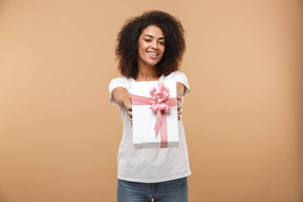 Portrait of a cheerful young african woman giving present box isolated over beige background - Foto, Bild