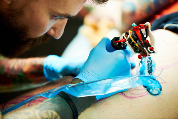 concentrated man tattoo master making tattoo on leg of client by tattoo machine, close-up - 写真・画像