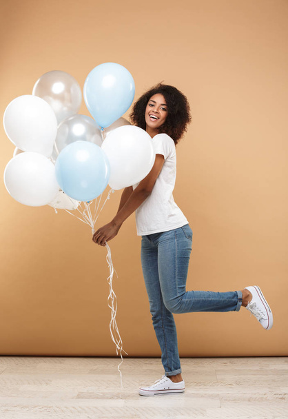 Full length portrait of a happy young african woman holding bunch of air balloons isolated over beige background - Zdjęcie, obraz