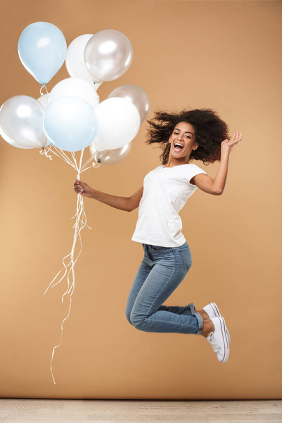 Full length portrait of a cheerful young african woman jumping while holding bunch of air balloons isolated over beige background - Zdjęcie, obraz