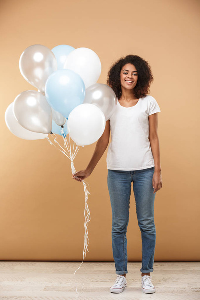 Full length portrait of a friendly young african woman holding bunch of air balloons isolated over beige background - Zdjęcie, obraz