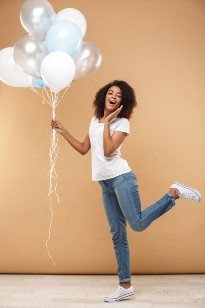 Full length portrait of a cheerful young african woman holding bunch of air balloons isolated over beige background - Fotografie, Obrázek