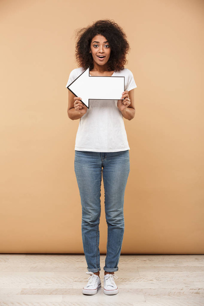 Full length portrait of a happy young african woman pointing away with an arrow isolated over beige background - Photo, Image