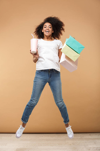 Full length portrait of a smiling young african woman jumping while holding shopping bags and drinking from a plastic cup isolated over beige background - Foto, immagini