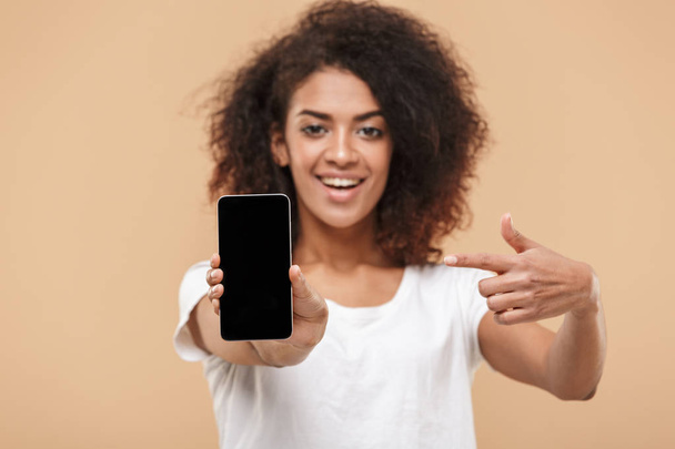 Portrait of a smiling young african woman poinitng finger at blank screen mobile phone isolated over beige background - Zdjęcie, obraz
