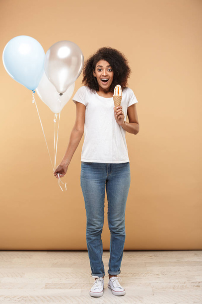 Full length portrait of a cheerful young african woman holding bunch of air balloons and an ice cream isolated over beige background - Фото, изображение