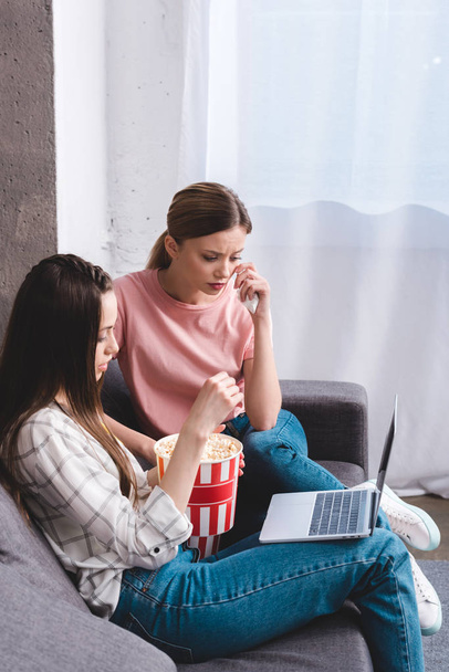 crying young woman watching movie with female friend eating popcorn and holding laptop on knees at home - Φωτογραφία, εικόνα