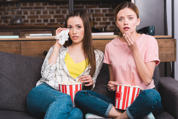 two upset female friends with buckets of popcorn watching movie on sofa at home  - Foto, immagini