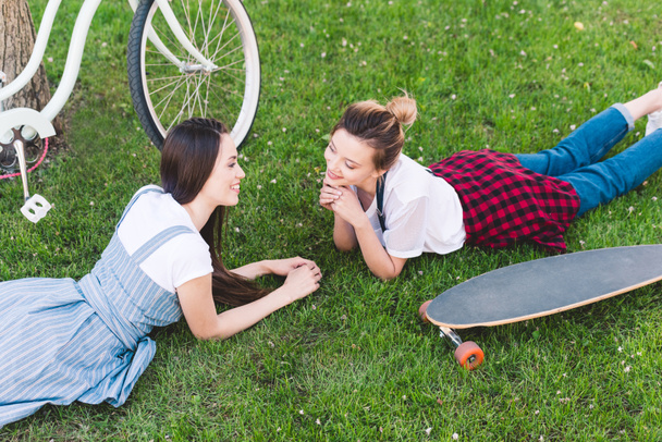 elevated view of smiling female friends laying on grass with bicycle and skateboard in park  - Fotó, kép