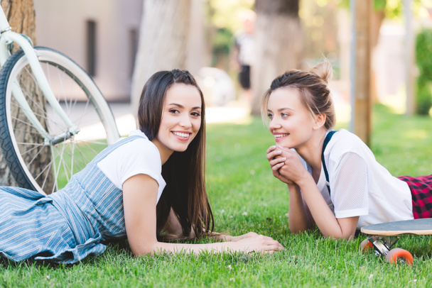 glimlachend vriendinnen opleggen van gras met fiets- en skateboard in park  - Foto, afbeelding