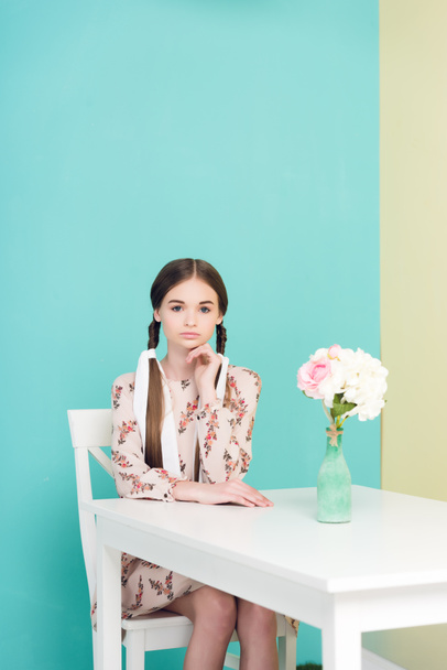menina adolescente atraente com tranças no vestido de verão sentado à mesa com flores, em azul
 - Foto, Imagem