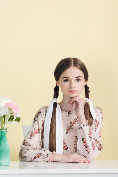 beautiful youth girl with braids sitting at table with flowers, isolated on yellow - Foto, Imagem