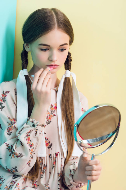 bella ragazza adolescente con trecce applicando rossetto con specchio
  - Foto, immagini