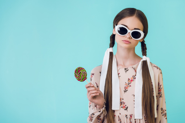 fashionable teen girl in sunglasses with braids holding lollipop, isolated on blue - Foto, afbeelding