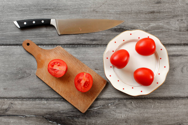 Table top view on three tomatoes on white porcelain plate with red dots, cutting board with tomato cut to half next to it, chef knife also laying on the gray wood desk. - Photo, image