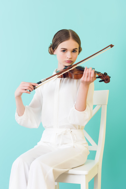teen girl playing violin and sitting on chair, isolated on turquoise  - Фото, зображення