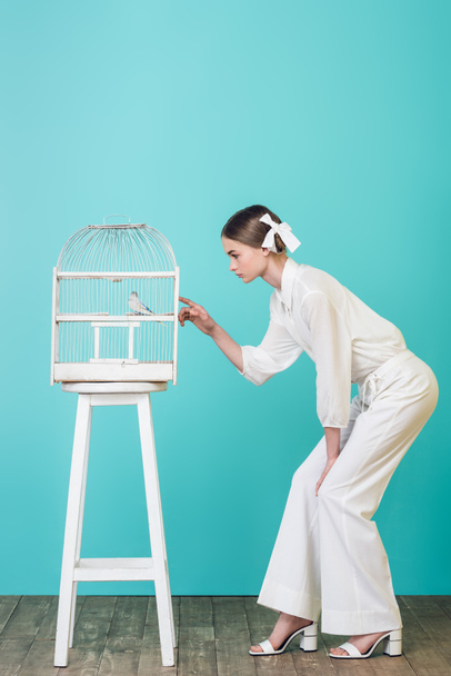 beautiful girl in white outfit looking parrot in cage, on turquoise - Foto, Imagen