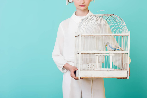 cropped view of girl in white holding cage with parrot, isolated on turquoise - Fotografie, Obrázek