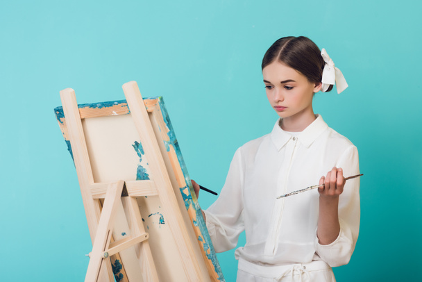 beautiful girl painting on easel with brush and palette, isolated on turquoise - Foto, immagini